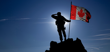 The backlit silhouette of a soldier standing on the top of a mountain, holding a Canadian flag hoisted on a flagpole.