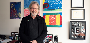 A man stands in an office before a wall of colourful children’s paintings. 
