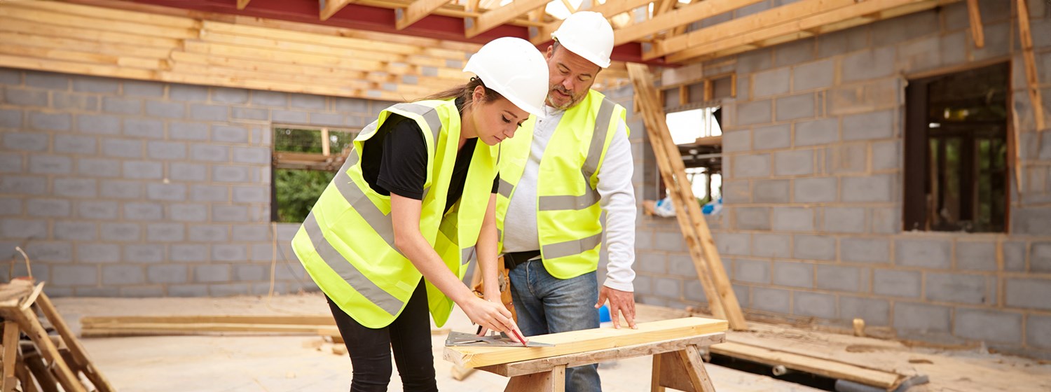 A woman and a man, wearing yellow reflective vests and white construction hats, lean over a woodworking bench on a construction site.