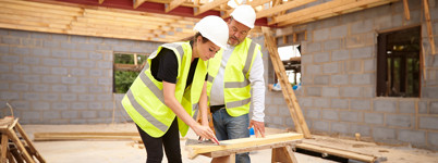 A woman and a man, wearing yellow reflective vests and white construction hats, lean over a woodworking bench on a construction site.