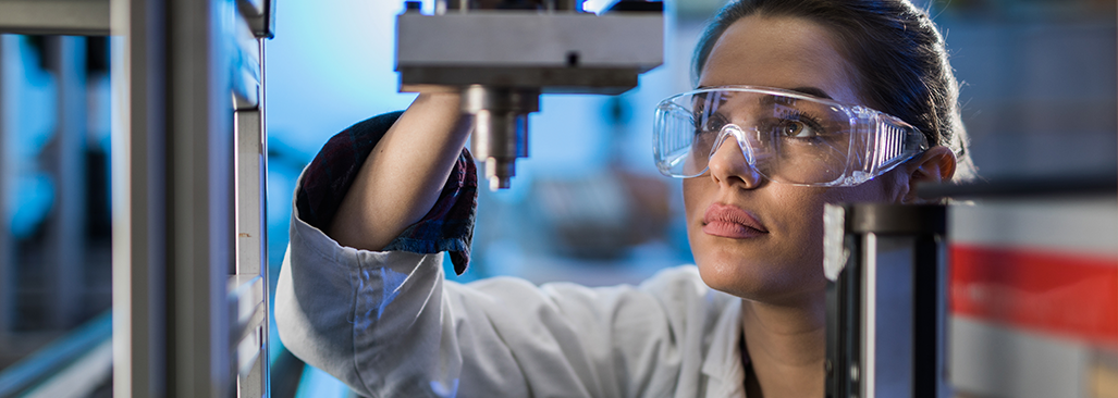 A woman wearing transparent safety glasses and a white lab coat examines a machine part on a manufacturing production line.