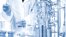 Gloved hands mix liquid in a container at a laboratory table covered in flasks and machinery. 