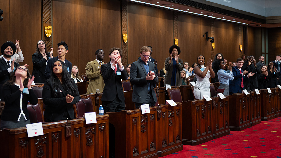 Young adults standing at wooden desks in the Senate Chamber clapping while looking upwards