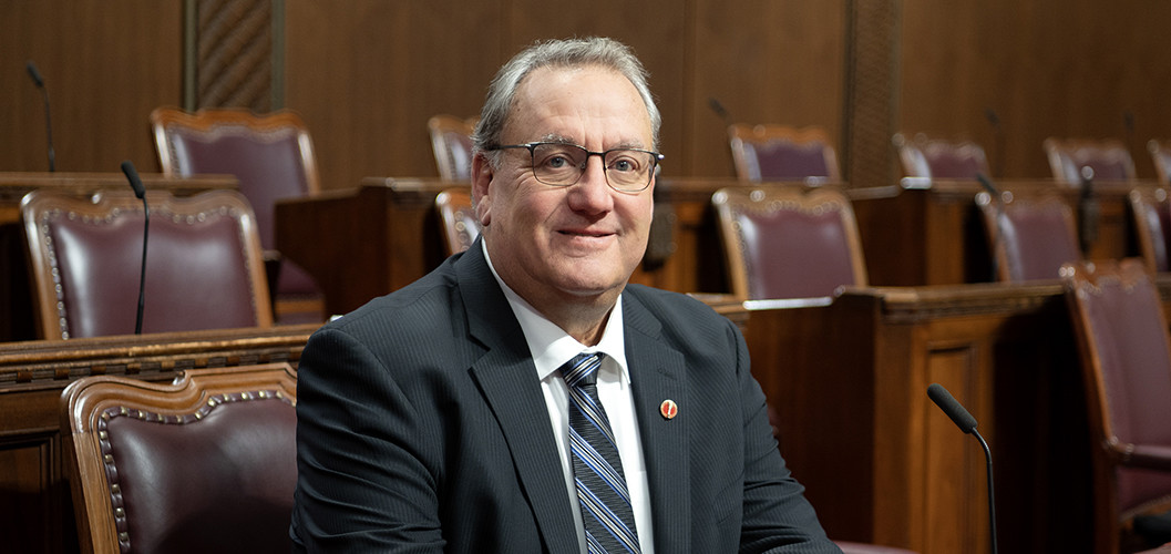 A man in a suit sitting at a wooden desk in the Senate Chamber.