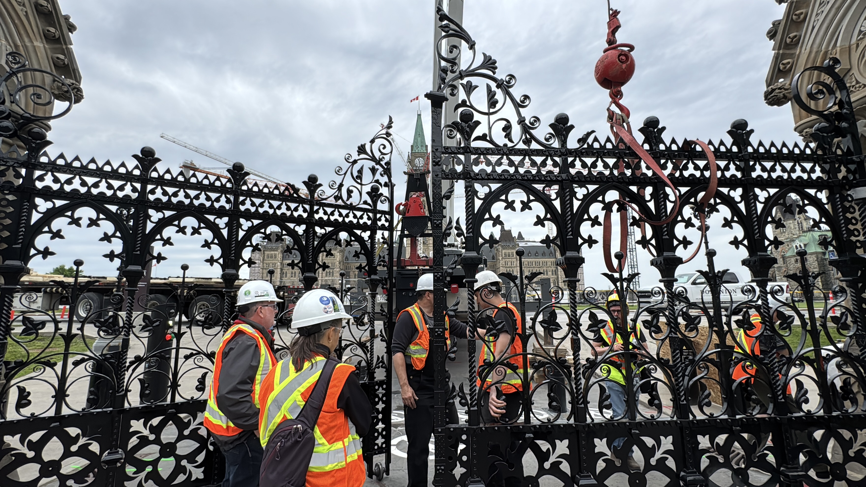A pair of large, black, partially opened iron gates in front of Centre Block on Parliament Hill, surrounded by people wearing personal protective equipment.