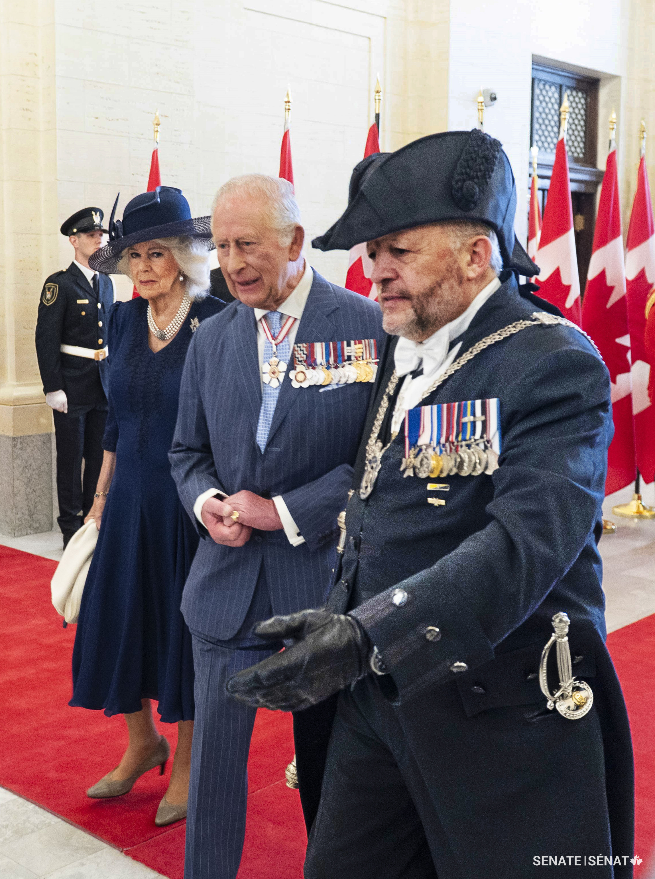 Usher of the Black Rod J. Greg Peters escorts the royal couple through the foyer of the Senate of Canada Building. The Usher is the monarch’s personal attendant in Parliament. His ceremonial sword — a gift from the King in March 2025 that bears the royal cypher on the guard — is visible at his side.