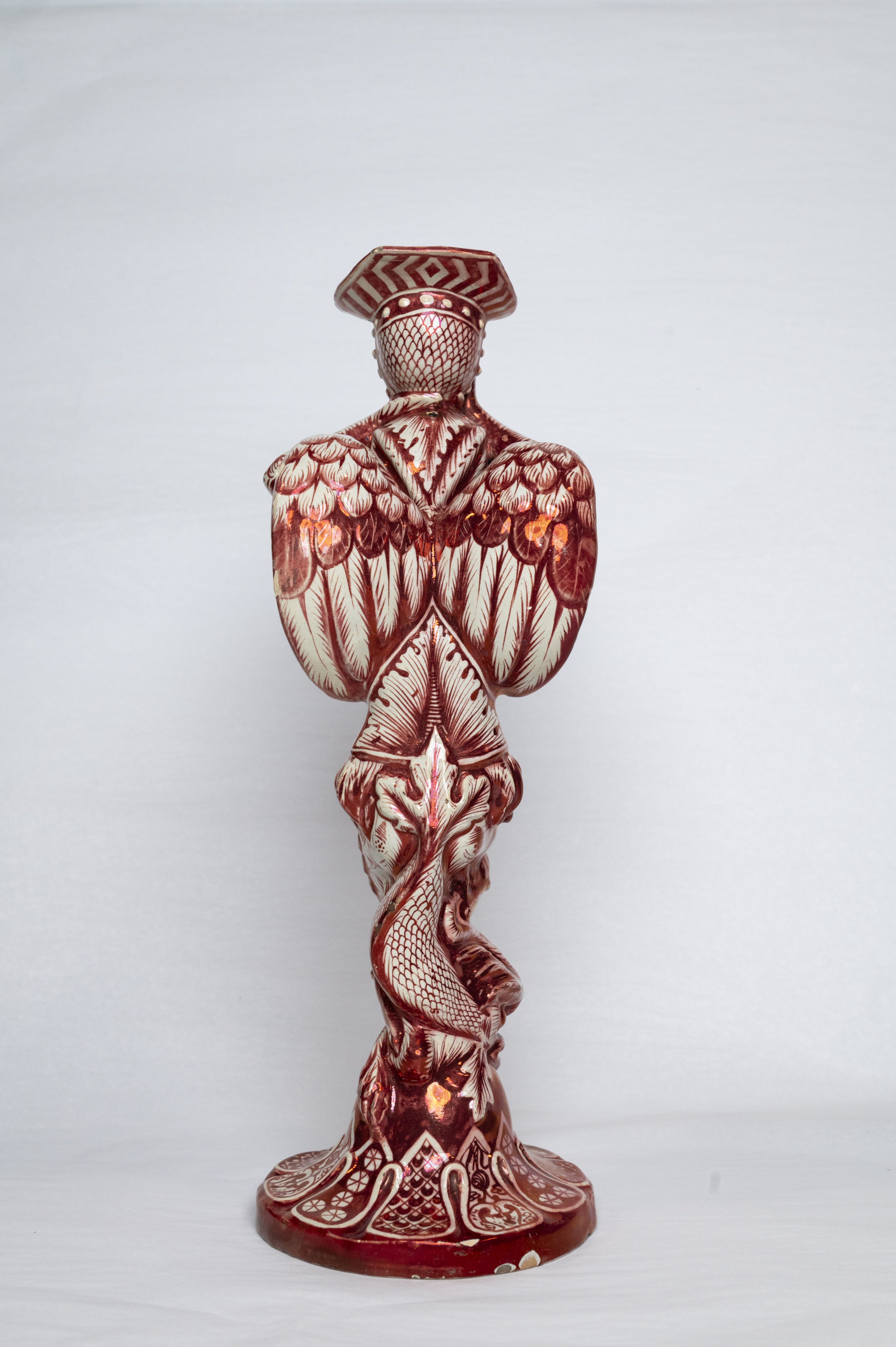 Le dos d’un bougeoir en céramique émaillée représentant une grotesque et où figurent, de haut en bas, un casque renversé, une paire d’ailes et une queue de dauphin.