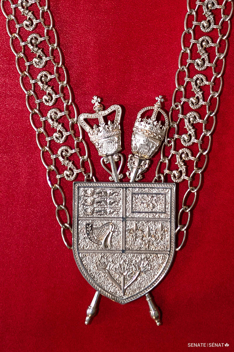 The shield from the Arms of Canada hangs at the back. It is superimposed over the maces of the Senate and the House of Commons, alluding to the fact that the Usher is the only senior officer of Parliament permitted on the floor of both chambers.