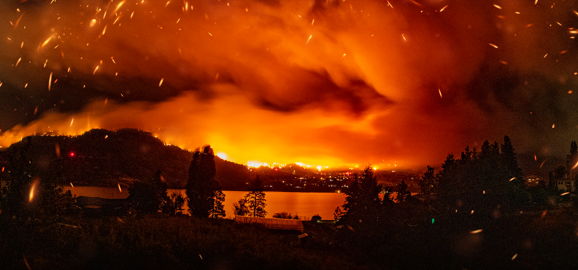 un incendie intense se propageant sur une colline et dans le paysage environnant, avec des flammes orange vif, une épaisse fumée rouge orangé et des braises lumineuses remplissant le ciel