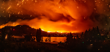 an intense wildfire burning across a hillside and surrounding landscape, with bright orange flames, thick red-orange smoke and glowing embers filling the sky