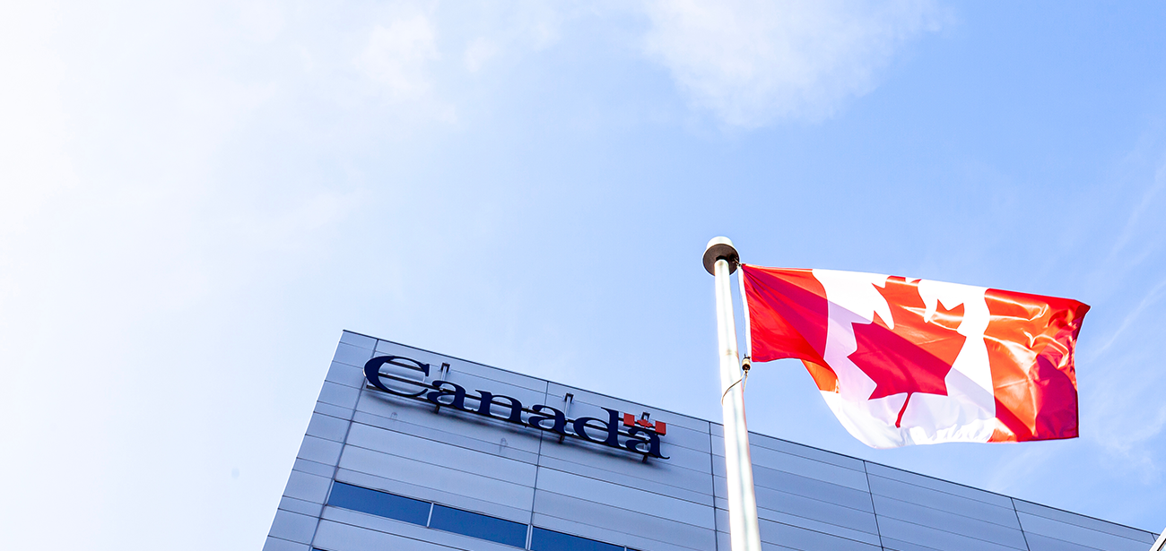 Canada’s national flag on a flagpole in front of a building bearing a Government of Canada sign, with a blue sky in the background.