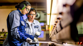 Two women dressed in safety gear use a press brake machine in a manufacturing facility.