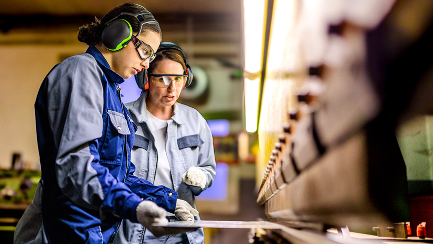 Two women dressed in safety gear use a press brake machine in a manufacturing facility.