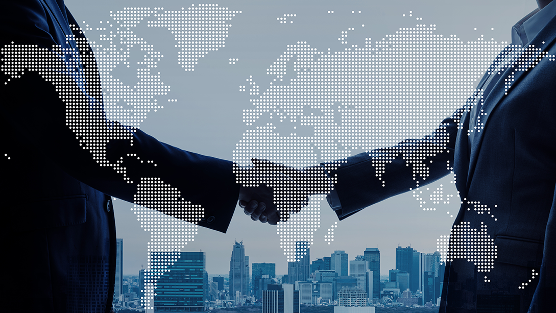 Layered pictures of a metropolitan skyline, an outlined world map and the silhouettes of two people in suits shaking hands.