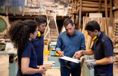A woman and three men review paperwork on a clipboard in a workshop.