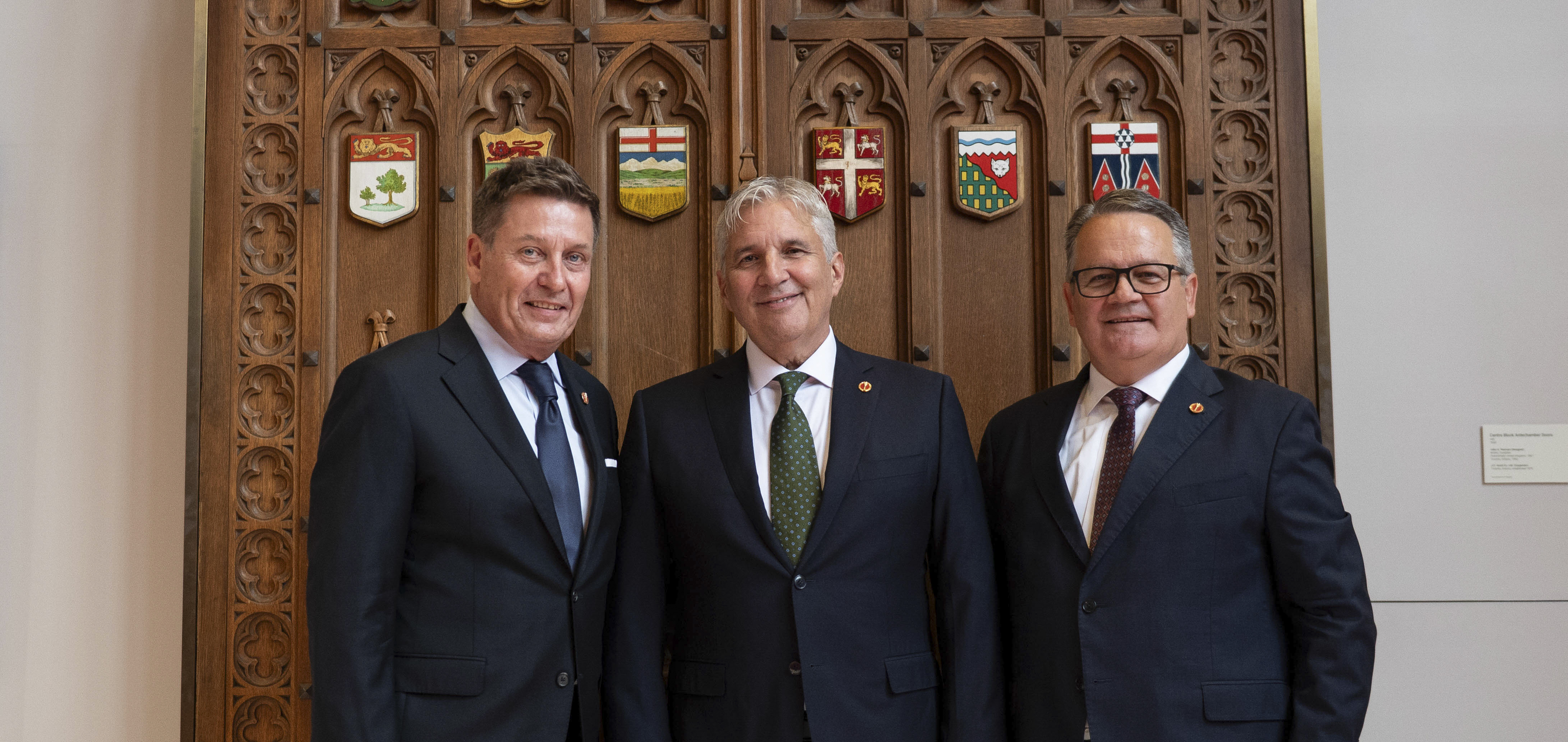 Trois hommes en costume devant des portes en bois ornées de plaques représentant les provinces et territoires du Canada.