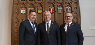 Three men wearing suits in front of wooden doors with plaques of Canada’s provinces and territories.