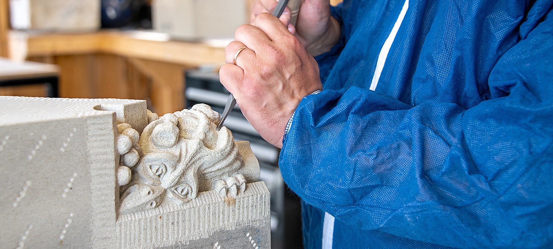 A close-up of a sculptor’s hands holding a chisel and carving a sculpture of a dragon with a bird’s beak.