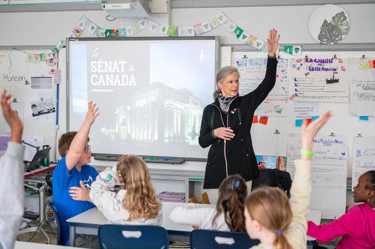 Une femme debout dans une salle de classe devant un écran affichant « Le Sénat du Canada », tandis que des élèves assis à leur pupitre lèvent la main.