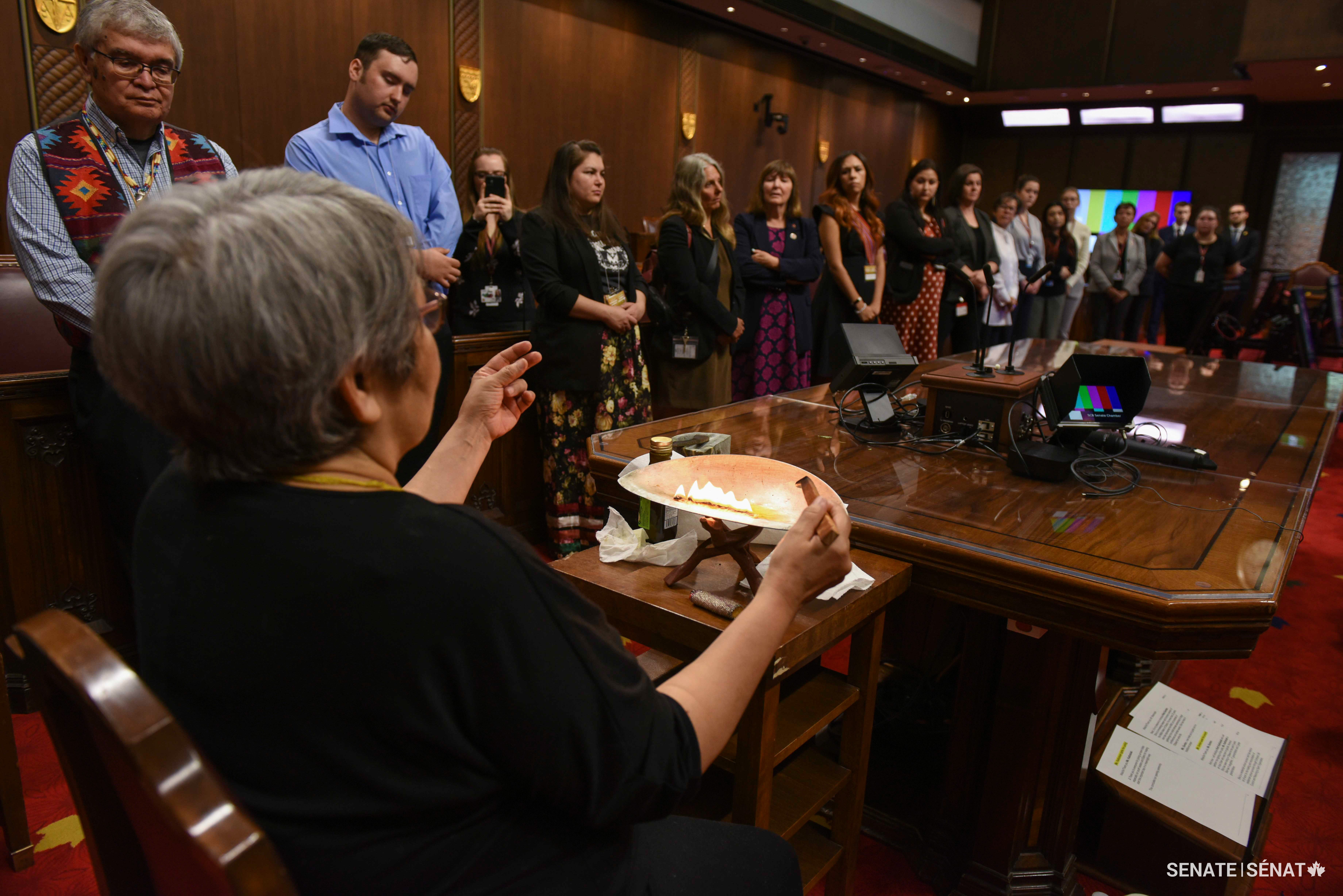 L’aînée inuite Ruth Kadlutsiak, assise à la table du greffier, allume un « qulliq » et en explique l’histoire et l’utilisation devant les participants de Voix de jeunes leaders autochtones, les sénateurs, le personnel du Sénat et d’autres invités dans la Chambre rouge.